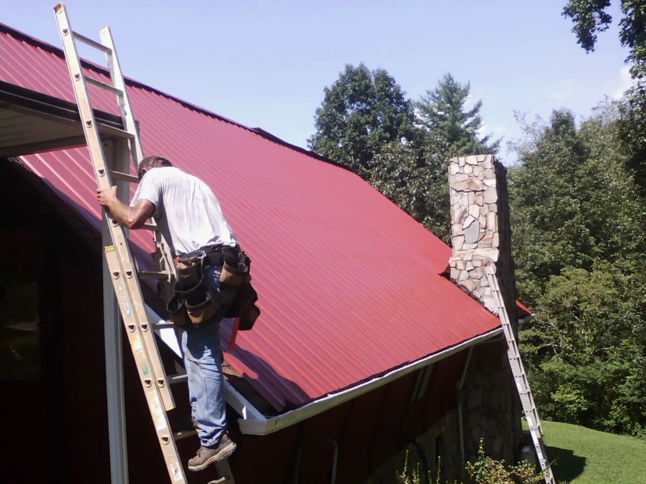 Roofer on a ladder beside a red metal roof and stone chimney — Soffit & Fascia Repair by Roofing Brothers in Riggins